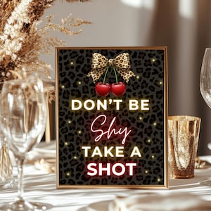 以下が含まれることがあります： ヒョウ柄の背景に「DON'T BE SHY TAKE A SHOT」のネオン風の文字が書かれた額入りのサイン。サインの上部にはリボンと2つのさくらんぼが描かれています。サインは、グラスと金色のカップが置かれたテーブルの上にあります。