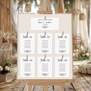 Puede incluir: Un plano de asientos de boda en un caballete. El gráfico presenta seis asignaciones de mesa, cada una con nombres de invitados, y un encabezado que dice "FIND YOUR TABLE Olivia & Jackson 12.10.2016". El diseño incluye detalles florales y se sujeta con pinzas de madera.