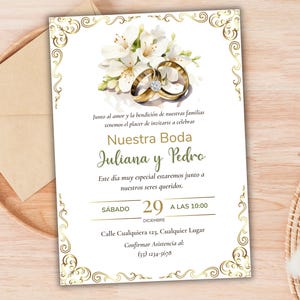 Op de afbeelding: Een rechthoekige trouwkaart met een gouden filigraanrand, een aquarelillustratie van witte bloemen en twee gouden trouwringen met een diamant. De tekst luidt: Nuestra Boda, Juliana y Pedro, Sábado 29 a las 10:00.