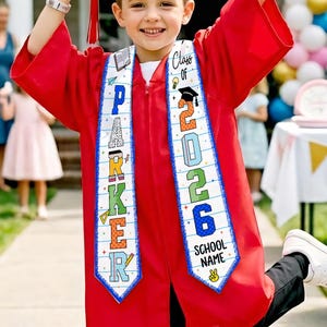 May include: A child in a red graduation gown and cap, holding a diploma. The sash reads "Class of 2026" and "School Name". A banner in the background says "Congratulations". The child is smiling and celebrating their graduation.