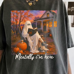 May include: A charcoal gray t-shirt featuring a graphic of a ghost sitting in a chair reading a book in front of a house with pumpkins nearby. The text "Mentally i'm here" is printed in white script below the illustration of the ghost.