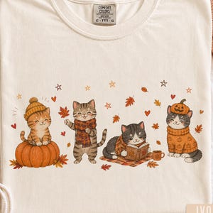 May include: An ivory t-shirt featuring a graphic of four cats in autumn attire. One cat sits on a pumpkin, one wears a plaid scarf, one reads a book on a blanket, and one wears a pumpkin hat. The design includes falling leaves, hearts, and stars.