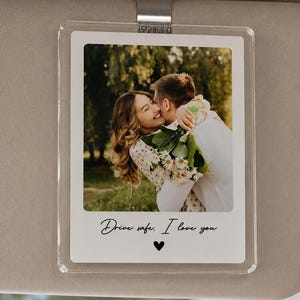 May include: A rectangular clear acrylic car visor clip featuring a printed photograph of a couple embracing outdoors. The bottom white border displays the black cursive text Drive safe. I love you with a small black heart icon centered below it.