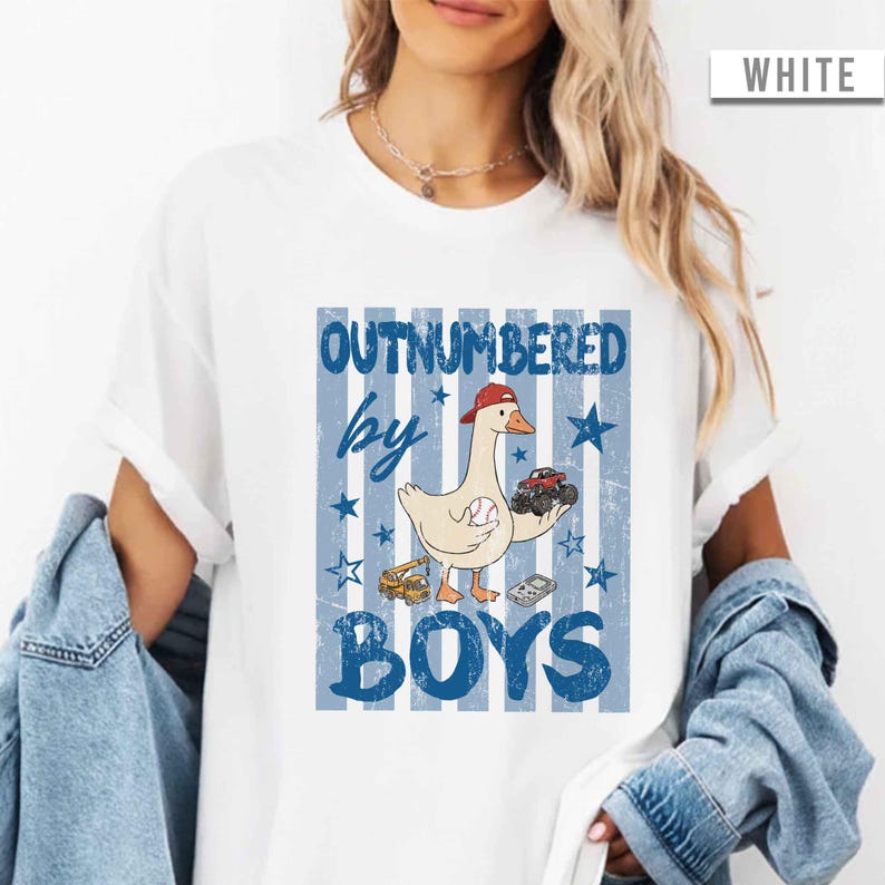 Peut inclure: T-shirt blanc avec un motif &agrave; rayures bleues et blanches. Le motif repr&eacute;sente une oie de dessin anim&eacute; portant une casquette rouge, tenant une balle de baseball et un monster truck. Le texte dit "OUTNUMBERED by BOYS."