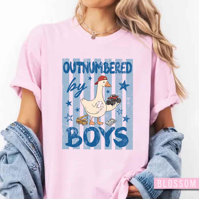 Peut inclure: T-shirt rose clair avec un motif graphique repr&eacute;sentant une oie portant une casquette rouge et tenant un camion jouet et une balle de baseball. Le texte "OUTNUMBERED by BOYS" est en bleu. Le fond pr&eacute;sente des rayures et des &eacute;toiles bleues.