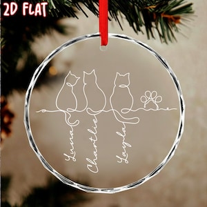 K&ouml;nnte beinhalten: Ein klarer, runder Weihnachtsschmuck mit rotem Band. Das Ornament zeigt eine Strichzeichnung von drei Katzen und einem Pfotenabdruck. Die Namen "Luna", "Charlie" und "Layla" sind darunter geschrieben.