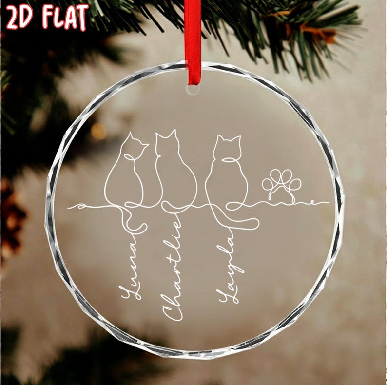 K&ouml;nnte beinhalten: Ein klarer, runder Weihnachtsschmuck mit rotem Band. Das Ornament zeigt eine Strichzeichnung von drei Katzen und einem Pfotenabdruck. Die Namen "Luna", "Charlie" und "Layla" sind darunter geschrieben.