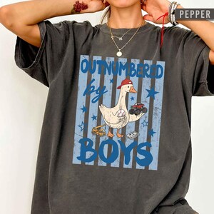 Peut inclure: T-shirt gris fonc&eacute; avec l'inscription bleue "OUTNUMBERED by BOYS". Un dessin d'oie portant une casquette rouge et tenant une balle de baseball et un monster truck est au centre. Des &eacute;toiles et des rayures bleues compl&egrave;tent le design.