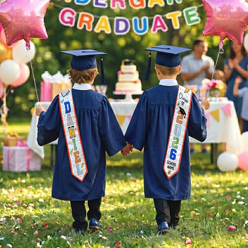 May include: Two children in navy blue graduation gowns and caps hold hands outdoors. Each child wears a sash that says "Class of 2026" and "School Name". A banner reads "Kindergarten Graduate". Pink star-shaped balloons are in the background.