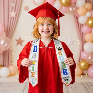 May include: A smiling child in a red graduation gown and cap, holding a white sash. The sash has the text "DANIEL" and "Class of 2026 SCHOOL NAME" in colorful letters. Balloons and stars decorate the background.