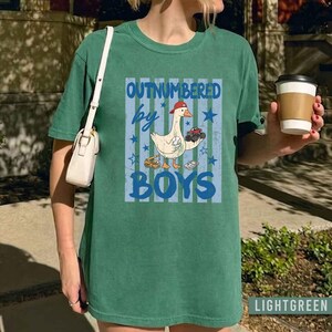 Peut inclure: T-shirt vert clair avec l'inscription "OUTNUMBERED by BOYS" en bleu. Le motif repr&eacute;sente une oie de dessin anim&eacute; portant une casquette rouge, tenant des voitures jouets et un engin de chantier. Le t-shirt a un aspect l&eacute;g&egrave;rement d&eacute;lav&eacute;.