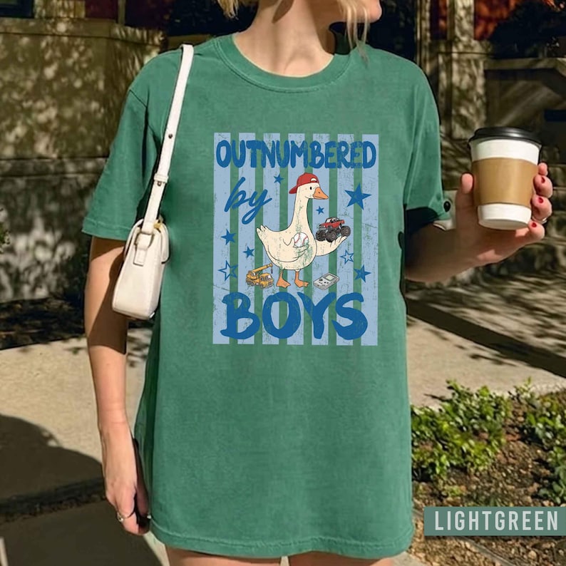 Peut inclure: T-shirt vert clair avec l'inscription "OUTNUMBERED by BOYS" en bleu. Le motif repr&eacute;sente une oie de dessin anim&eacute; portant une casquette rouge, tenant des voitures jouets et un engin de chantier. Le t-shirt a un aspect l&eacute;g&egrave;rement d&eacute;lav&eacute;.
