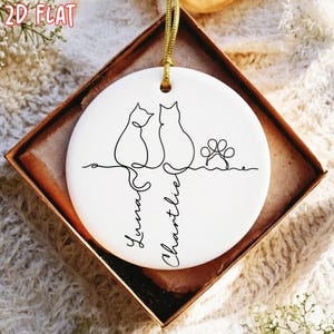 K&ouml;nnte beinhalten: Runder wei&szlig;er Keramikschmuck mit goldener Aufh&auml;ngeschnur. Er zeigt eine schwarze Strichzeichnung von zwei Katzen, einen Pfotenabdruck und die Namen "Luna" und "Charlie" in Schreibschrift. Der Text "2D FLAT" befindet sich in der oberen linken Ecke.