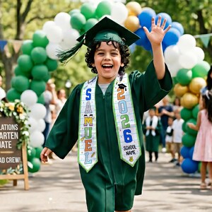May include: A young child in a green graduation gown and cap, waving with a big smile. The sash reads "SAMUEL 2026 SCHOOL NAME". Balloons in green, white, and blue create an archway in the background. A sign on the left reads "CONGRATULATIONS KINDERGARTEN GRADUATE".