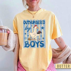 Peut inclure: T-shirt jaune beurre avec l'inscription "OUTNUMBERED by BOYS". Le motif repr&eacute;sente une oie de dessin anim&eacute; portant une casquette rouge et tenant des voitures jouets, avec des &eacute;toiles et des rayures bleues.