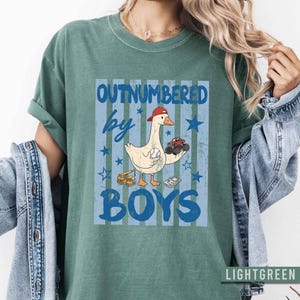 Peut inclure: T-shirt vert clair avec un motif &laquo;&nbsp;OUTNUMBERED by BOYS&nbsp;&raquo;. Le motif repr&eacute;sente une oie de dessin anim&eacute; portant une casquette rouge, tenant une balle de baseball et un camion jouet. Des &eacute;toiles et des rayures bleues compl&egrave;tent le motif.