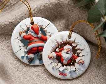 Puede incluir: Dos adornos redondos de cerámica blanca con cordones dorados. Uno muestra a Santa Claus desde atrás gateando a través de una pared agrietada. El otro muestra a Santa de frente, emergiendo de una pared agrietada con el texto rojo 2026 debajo.