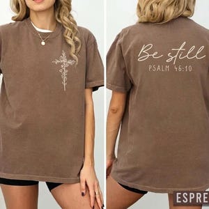 May include: Espresso-colored t-shirt with a white floral cross design on the front. The back of the shirt features the text "Be still" and "PSALM 46:10" in a cursive font. The shirt is a comfortable, casual style.