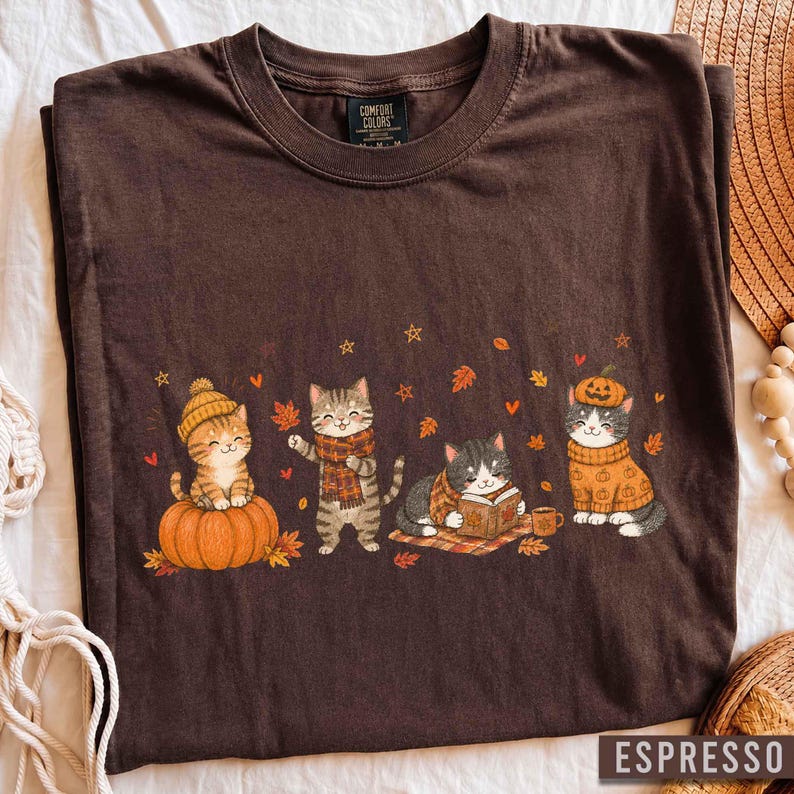 May include: A dark brown t-shirt featuring a graphic of four cats in autumn attire. One cat sits on a pumpkin, one wears a scarf, one reads a book, and one wears a pumpkin hat. The Comfort Colors brand label is visible at the inner neckline.