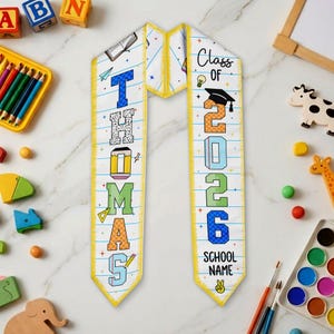 May include: A graduation sash with the name "Thomas" and the year "2026" printed on it. The sash is white with a yellow border and features colorful text and graphics, including a graduation cap and pencil.