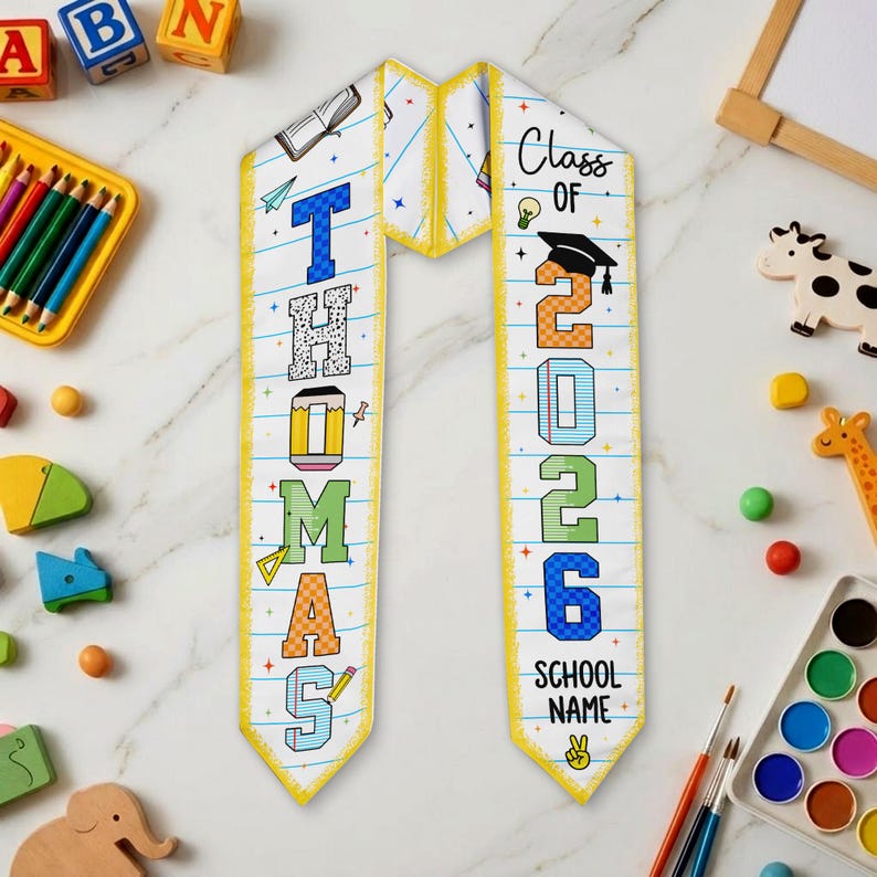 May include: A graduation sash with the name "Thomas" and the year "2026" printed on it. The sash is white with a yellow border and features colorful text and graphics, including a graduation cap and pencil.