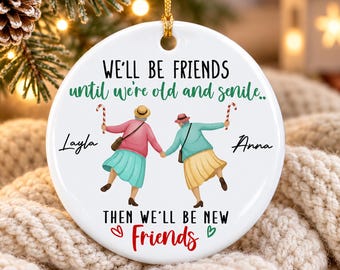 May include: A round white ceramic ornament featuring an illustration of two elderly figures holding candy canes. The text reads: WE'LL BE FRIENDS until we're old and senile.. Layla, Anna, THEN WE'LL BE NEW Friends. It includes a gold hanging cord.