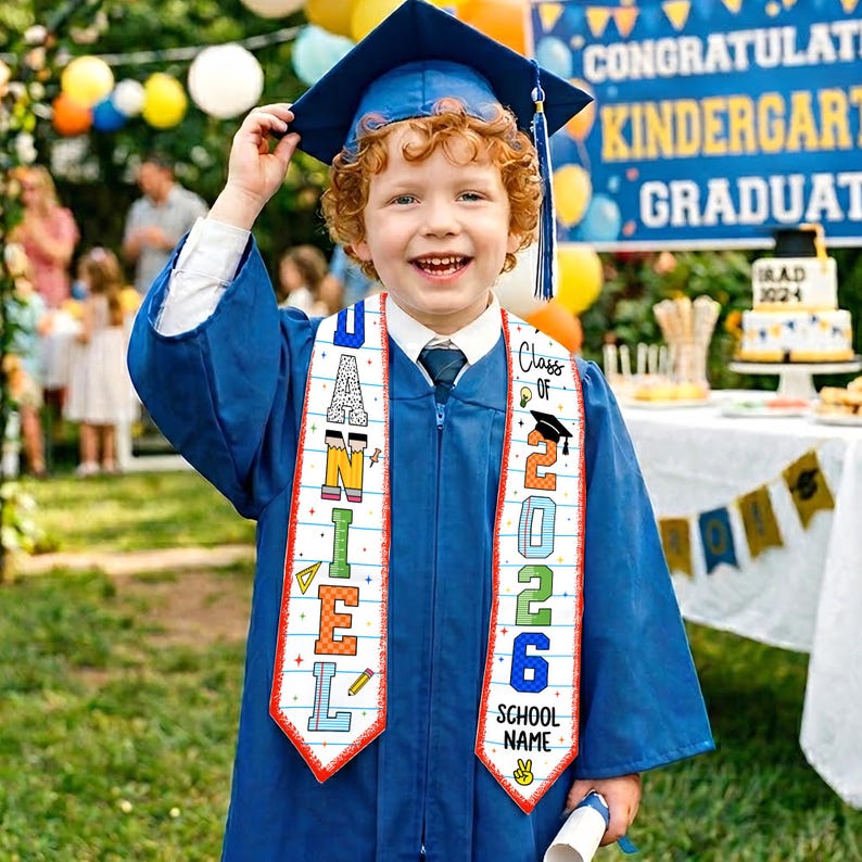 May include: A young child in a blue graduation gown and cap smiles, holding the cap. The sash reads "DANIEL" and "Class of 2026." A banner in the background reads "Congratulations Kindergarten Graduate."