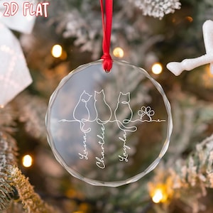 K&ouml;nnte beinhalten: Ein klarer, runder Weihnachtsschmuck mit rotem Band. Der Schmuck zeigt drei Strichzeichnungen von Katzen, einen Pfotenabdruck und die Namen "Luna", "Charlie" und "Layla" in Schreibschrift. Der Schmuck h&auml;ngt vor einem Weihnachtsbaum.