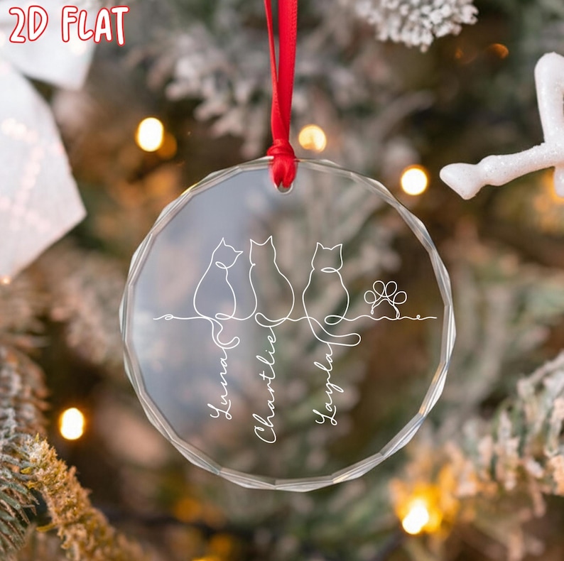 K&ouml;nnte beinhalten: Ein klarer, runder Weihnachtsschmuck mit rotem Band. Der Schmuck zeigt drei Strichzeichnungen von Katzen, einen Pfotenabdruck und die Namen "Luna", "Charlie" und "Layla" in Schreibschrift. Der Schmuck h&auml;ngt vor einem Weihnachtsbaum.
