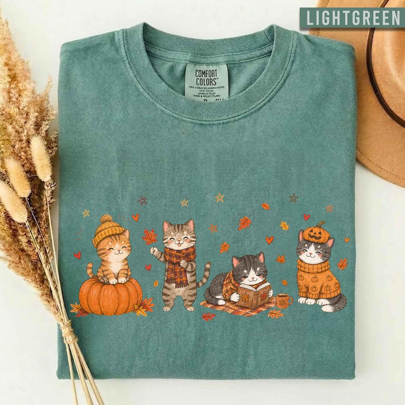 May include: A sage green t-shirt featuring a graphic of four cats in autumn attire. One cat sits on a pumpkin, one wears a plaid scarf, one reads a book, and one wears a pumpkin hat. The design includes scattered orange leaves and stars.