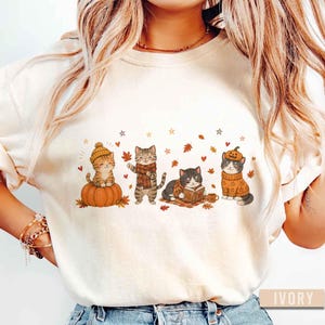 May include: An ivory-colored t-shirt featuring a graphic print of four cats in autumn-themed attire. One cat sits on a pumpkin, another wears a scarf, one reads a book, and the last wears a pumpkin hat. The design includes falling leaves and stars.