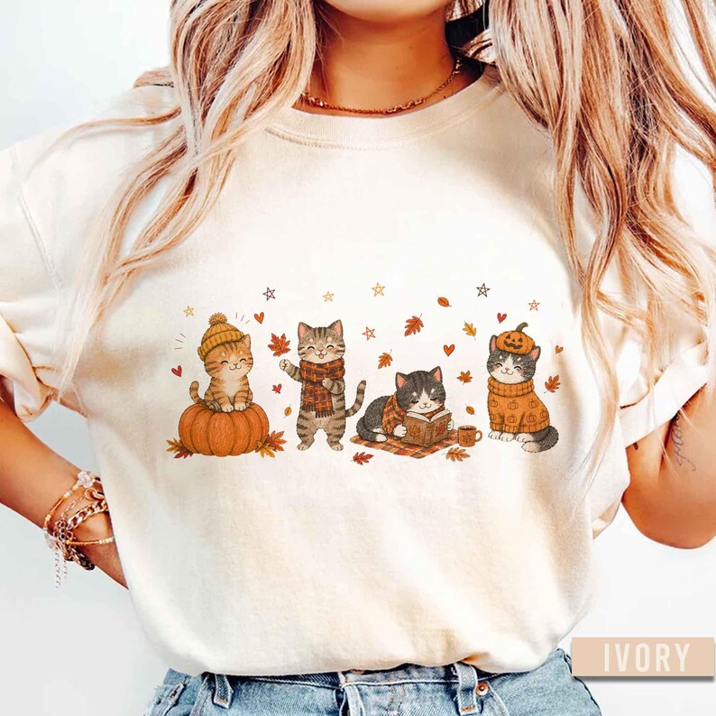 May include: An ivory-colored t-shirt featuring a graphic print of four cats in autumn-themed attire. One cat sits on a pumpkin, another wears a scarf, one reads a book, and the last wears a pumpkin hat. The design includes falling leaves and stars.