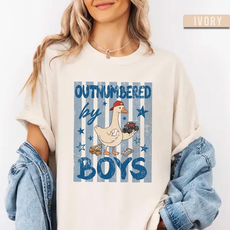 Peut inclure: T-shirt ivoire avec un motif &laquo; OUTNUMBERED by BOYS &raquo;. Le motif repr&eacute;sente une oie de dessin anim&eacute; portant une casquette rouge et tenant une balle de baseball et un camion jouet. Des &eacute;toiles et des rayures bleues compl&egrave;tent le design.
