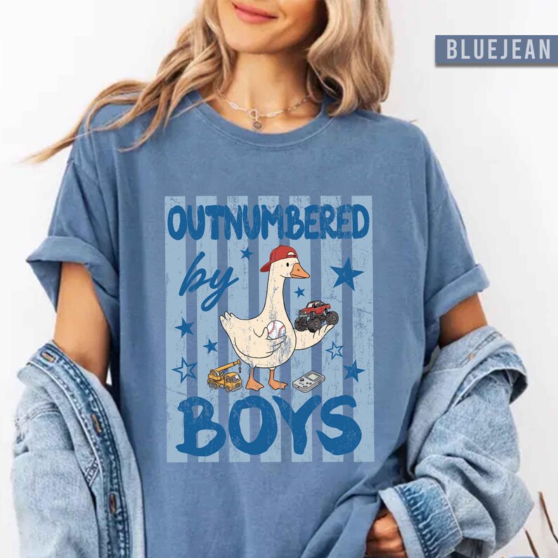 Peut inclure: T-shirt bleu avec l'inscription "OUTNUMBERED by BOYS" et une oie de dessin anim&eacute; portant une casquette de baseball, tenant une voiture jouet et une balle de baseball. Le design comprend des &eacute;toiles et des v&eacute;hicules jouets.