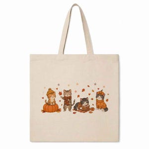 May include: A beige canvas tote bag featuring a printed illustration of four cats in autumn-themed attire. One cat sits on a pumpkin, another wears a scarf, one reads a book, and the last wears a pumpkin hat, all surrounded by falling leaves.