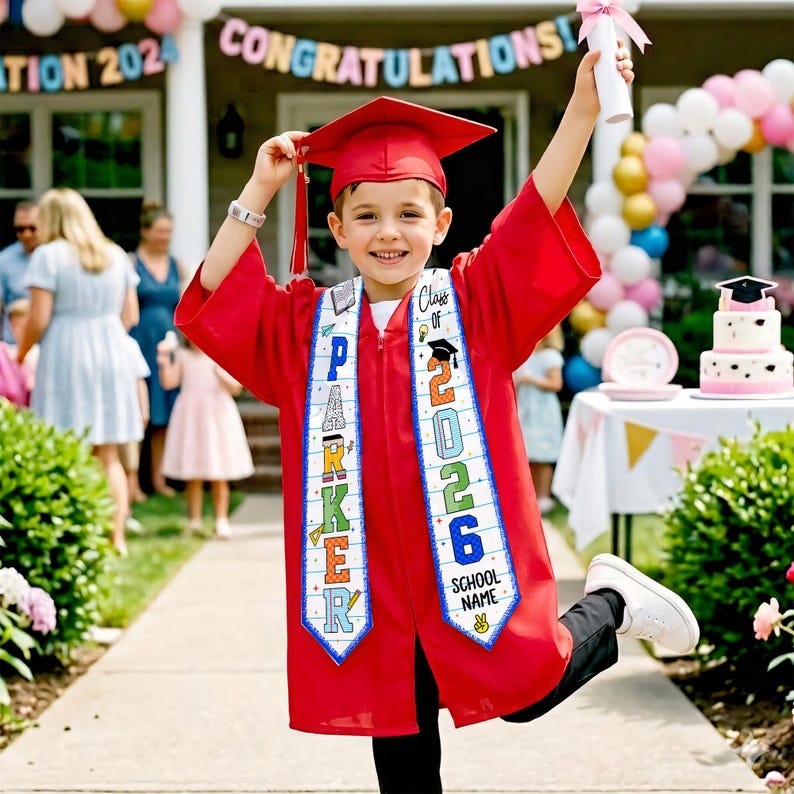 May include: A child in a red graduation gown and cap, holding a diploma. The sash reads "Class of 2026" and "School Name". A banner in the background says "Congratulations". The child is smiling and celebrating their graduation.