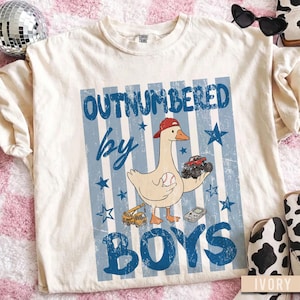 Peut inclure: T-shirt de couleur ivoire avec un motif &agrave; rayures bleues et blanches. Le t-shirt pr&eacute;sente une oie de dessin anim&eacute; portant une casquette rouge, tenant une balle de baseball et un monster truck. Le texte dit "OUTNUMBERED by BOYS."