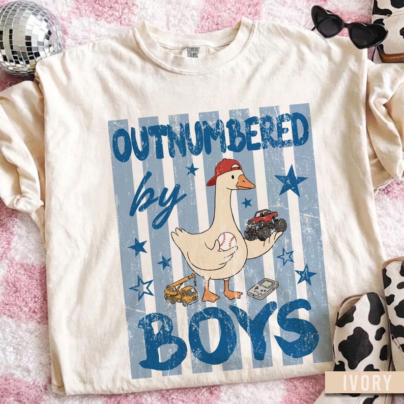 Peut inclure: T-shirt de couleur ivoire avec un motif &agrave; rayures bleues et blanches. Le t-shirt pr&eacute;sente une oie de dessin anim&eacute; portant une casquette rouge, tenant une balle de baseball et un monster truck. Le texte dit "OUTNUMBERED by BOYS."