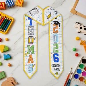 May include: A personalized graduation sash with the name "THOMAS" and the year "2026". The sash is white with colorful block letters, a yellow border, and school-themed illustrations. The words "School Name" are also visible.