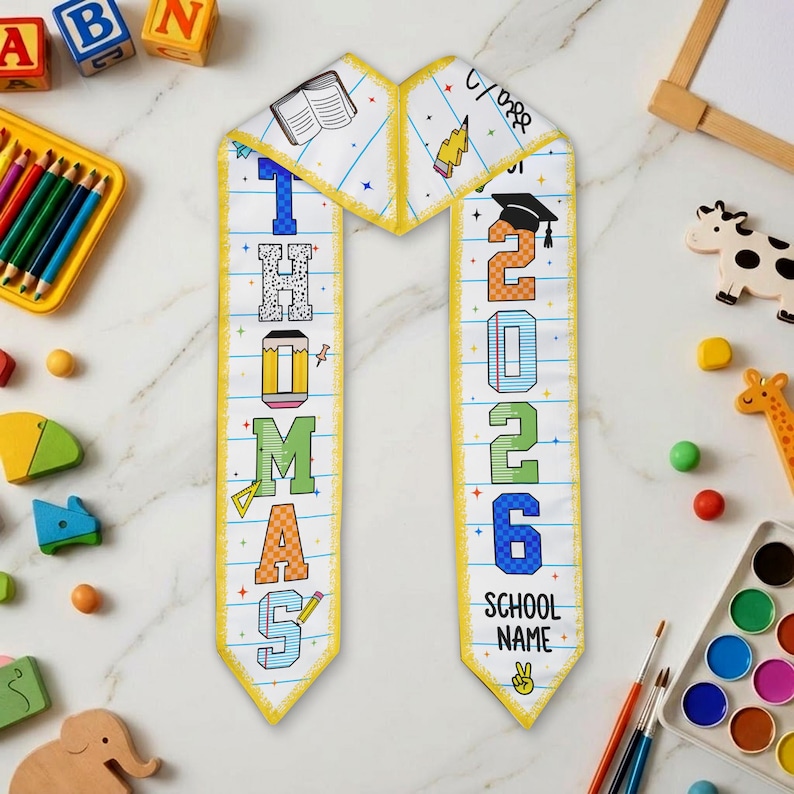May include: A personalized graduation sash with the name "THOMAS" and the year "2026". The sash is white with colorful block letters, a yellow border, and school-themed illustrations. The words "School Name" are also visible.