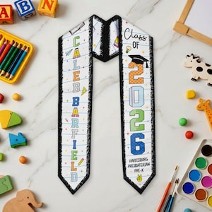 May include: White graduation sash with black trim. The sash features the name "CALEB GARFIELD" in colorful letters on one side and "Class of 2026" on the other. The sash also includes the school name "Harrisburg Presbyterian Pre-K".