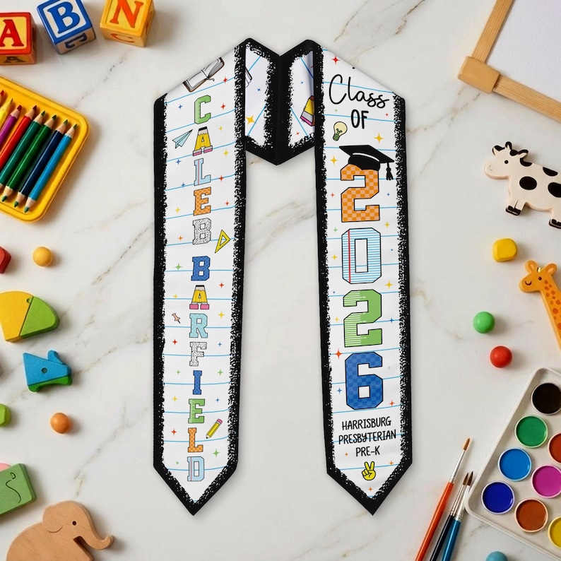 May include: White graduation sash with black trim. The sash features the name "CALEB GARFIELD" in colorful letters on one side and "Class of 2026" on the other. The sash also includes the school name "Harrisburg Presbyterian Pre-K".