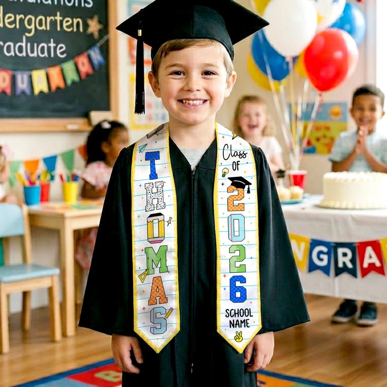 May include: A young child in a black graduation gown and cap smiles, wearing a personalized sash that reads "Thomas" and "Class of 2026." The sash is white with colorful lettering and illustrations. Balloons and a cake are in the background.