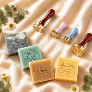 May include: Four square soap bars in gray, green, tan, and yellow, each featuring unique embossed designs and text including "Handmade by Jillian," "Handmade - WITH LOVE -," and "THE BOTANICAL CORNER." Four wax seal stamps with colored handles.