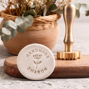 Könnte beinhalten: Ein runder, cremefarbener handgefertigter Stempel mit dem Namen "Sharon" und einem floralen Design. Ein goldfarbenes Stempelwerkzeug und ein Holzschneidebrett befinden sich im Hintergrund. Ein geflochtener Korb mit Trockenblumen und Grünzeug ist ebenfalls zu sehen.