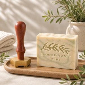Puede incluir: Una pastilla de jabón de hierbas hecho a mano con un diseño de rama de olivo y el texto "Handmade Herbal Soap by Lisa" está sobre una bandeja de madera. Un sello con mango de madera y base dorada está junto al jabón.