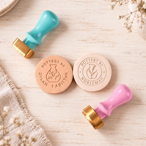 May include: Two metal stamps with turquoise and pink handles, each featuring a brass base. Beside them are two circular clay impressions. One impression reads POTTERY BY DYLAN TABITHA with a vase icon, and the other reads POTTERY BY NOBLEMAN.