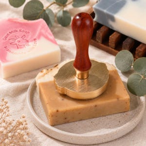 May include: A brass soap stamp with a dark brown wooden handle rests on a rectangular tan bar of soap. Nearby, a pink and white bar of soap features a goat head graphic and the text Goat Milk Soap Handmade with Love, alongside a white bar.