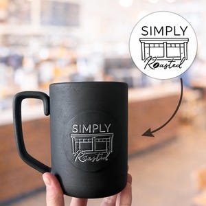 Puede incluir: Una taza de cerámica negra con un asa curvada. La taza presenta un emblema circular con las palabras "SIMPLY Roasted" y un gráfico de una fachada de tienda. La taza está sujeta por una mano.