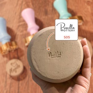 May include: A beige pottery bowl with the text "Norris Nook Pottery" and "Priscilla Pottery S05". Pastel-coloured pottery stamps are in the background. The bowl is held in a person's hand.